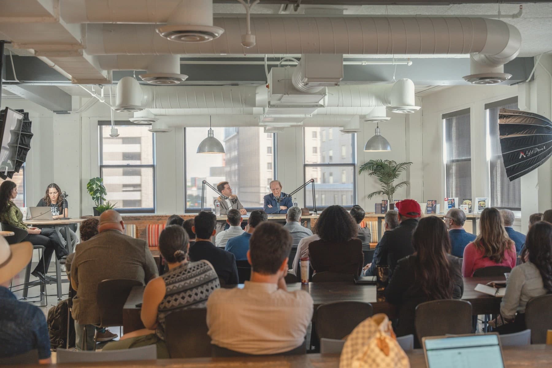 The third floor from the back: pendant lamps, exposed ductwork, tall windows onto downtown San Antonio, and a full audience at long tables facing a panel.