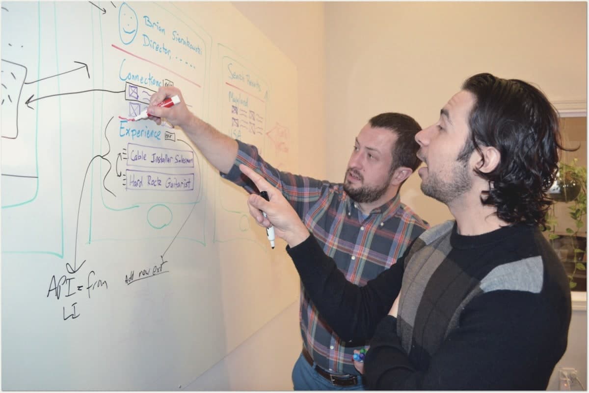 Brian Sierakowski at a whiteboard with a founder, sketching a product wireframe while the founder points at the screen layout.